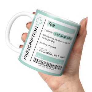 May include: A light blue ceramic mug with a white handle. The mug has a prescription label that reads "Tea" and includes the text "Patient: ANY NAME HERE" and "One dose to be taken orally 3-4 times per day. Refill as required." The label also includes a handwritten signature that reads "T. Caddie" and the text "Dr. T. Caddie". The mug has a barcode and the text "1 dose" and "325ml".