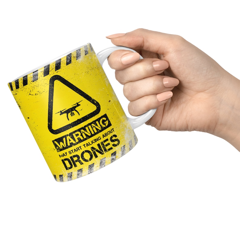 May include: A white ceramic mug with a yellow and black warning sign design. The sign reads "Warning May Start Talking About Drones".