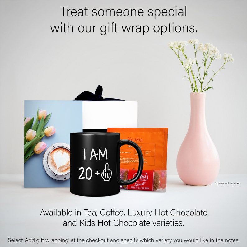 May include: A black mug with the text "I AM 20+" and a middle finger emoji. The mug is next to a box wrapped in white paper and a packet of hot chocolate mix. The background is a light gray wall with a vase of white flowers.