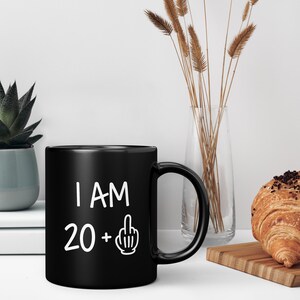 May include: Black coffee mug with white text that reads "I AM 20 +" with a middle finger emoji.