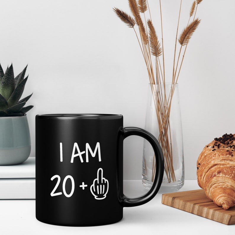 May include: Black coffee mug with white text that reads "I AM 20 +" with a middle finger emoji.