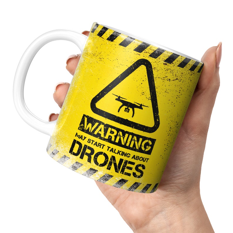 May include: A yellow mug with a black warning sign that says "Warning May Start Talking About Drones". The sign has a black triangle with a drone inside. The mug has a white handle.