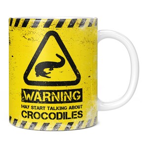 Könnte beinhalten: Eine gelbe Tasse mit einem schwarzen Warnschild, auf dem "WARNUNG KANN ÜBER KROKODILE REDEN" steht. Das Schild hat ein schwarzes Dreieck mit einem Krokodil darin. Die Tasse hat einen weißen Henkel.