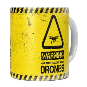 May include: A yellow mug with a black warning sign that says "Warning May Start Talking About Drones". The sign has a black triangle with a drone inside. The mug has a white handle.
