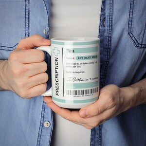 May include: White ceramic mug with a blue and white prescription label design. The label reads "PRESCRIPTION", "Tea", "Patient: ANY NAME HERE", "One dose to be taken orally 3-4 times per day", "Refill as required", "I Caddie", "Dr. T. Caddie", "1 dose", "3.25ml", "0009922-876720-ET".