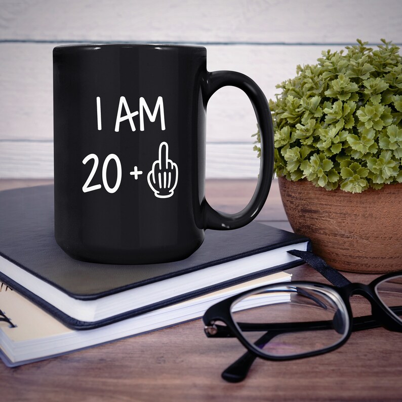 May include: Black coffee mug with white text that reads "I AM 20 +" with a middle finger emoji. The mug is sitting on a stack of notebooks and a wooden table.
