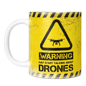 May include: A white ceramic mug with a yellow and black warning sign design. The sign features a drone icon and the text "WARNING MAY START TALKING ABOUT DRONES".