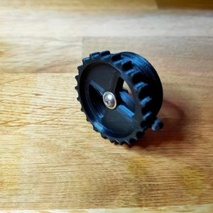 Könnte beinhalten: Ein schwarzes Kunststoffzahnrad mit einer silbernen Metallmitte. Das Zahnrad hat eine strukturierte Oberfläche und ist für den Einsatz in einer Maschine oder einem anderen mechanischen Gerät konzipiert.
