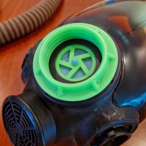 Op de afbeelding: Een zwart gasmasker met een felgroene filterring en een lichtblauw intern filter. Het masker heeft een ronde luchtinlaat en een geribbelde slang. De tekst "THE MASK IS NOT PART OF THE OFFER" is zichtbaar.