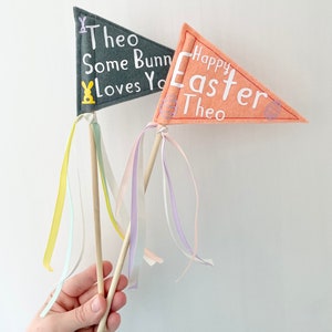 May include: Two felt pennants on wooden sticks with yellow, white and purple ribbons. The grey pennant says "Theo Some Bunny Loves You" with a yellow bunny and the orange pennant says "Happy Easter Theo" with a pink egg.