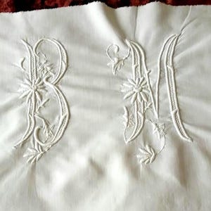 以下が含まれることがあります： 白いリネンの織物で、花柄のデザインで刺繍されたイニシャル「BM」が特徴です。イニシャルは装飾的な筆記体です。下端には、等間隔に配置された長方形の切り抜きが並んでいます。生地はわずかにしわがあります。