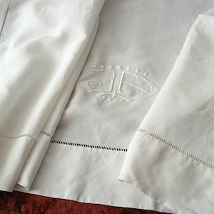 May include: White linen sheet with embroidered monogram. The sheet features a detailed, stylized monogram in the center, with decorative stitching along the edges. The fabric is folded, showing the texture and quality of the material.