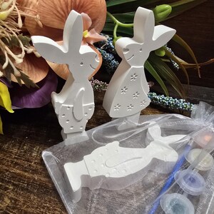 May include: White bunny figurines with cut-out details, standing upright, and a flat bunny figurine lying down. A small bag contains paint pots and a brush. The background includes flowers and greenery, suggesting a craft or Easter theme.