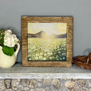 May include: A miniature scene featuring a framed landscape painting with a field of white flowers and mountains. A small white pitcher with white flowers and a woven basket are also on display. The painting has a rustic wooden frame.