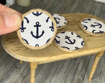 May include: A set of four round coasters with a nautical theme. Each coaster features a white fabric with a navy blue anchor pattern, framed by a natural jute rope edge. The coasters are displayed on a small wooden table.