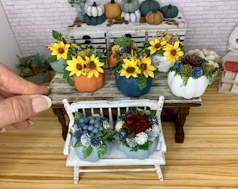 May include: A set of six miniature pumpkins decorated with fall-themed floral arrangements. The pumpkins are painted in shades of white, blue, and orange. The floral arrangements include sunflowers, succulents, and other fall foliage. The pumpkins are arranged on a white wooden bench and a wooden table.