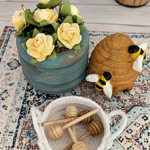 May include: A miniature scene featuring a blue wooden planter with yellow roses, a brown beehive with two bees, and a white rope basket filled with wooden honey dippers. The items are arranged on a patterned rug.