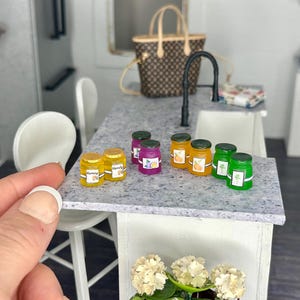 May include: A set of miniature glass jars filled with colorful jelly. The jars are arranged on a white countertop with a speckled pattern. The jars have labels with different fruit designs.