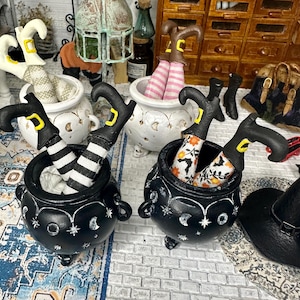 May include: Three black cauldron planters with white stars and moons painted on them. Each cauldron has a pair of witch legs sticking out of the top, wearing striped stockings and black shoes.