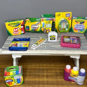 May include: A collection of Crayola art supplies, including crayons, glitter, markers, paint, and chalk, arranged on a small, weathered wooden table. The products are in various colorful packages, with the Crayola logo prominently displayed.