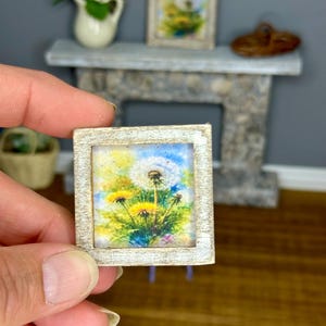 May include: Miniature framed artwork featuring a watercolor painting of dandelions. The painting has yellow, green, and blue hues, set against a white-washed wooden frame. The artwork is held by a person's hand, with a second similar piece in the background.