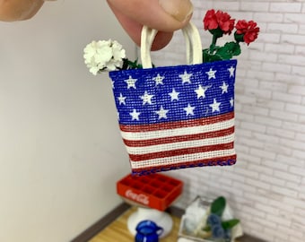 May include: Miniature tote bag with a patriotic design, featuring a blue field of white stars and red and white stripes. The bag has white handles and is filled with red and white flowers. The bag is held by a person's hand.