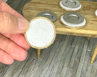 May include: Miniature dollhouse accessories. A white, round coaster with a woven jute border is held in a hand. Other coasters with metal plates and jute borders are on a wooden table. A small flower pot is in the background.