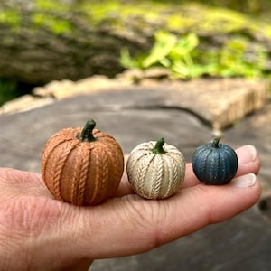 May include: Three miniature pumpkins with a textured cable knit pattern. The largest pumpkin is orange, the medium pumpkin is off-white, and the smallest pumpkin is dark blue. Each pumpkin features a small, dark green stem at the top.