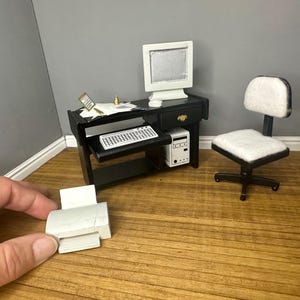 May include: Miniature black desk with a white computer monitor, keyboard, and computer tower. A white office chair and a small printer are also present. The desk has a drawer with a gold-colored handle. The scene is set on a wooden floor.