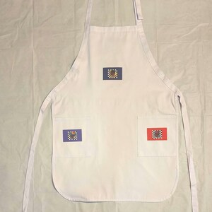 May include: White apron with three pockets. Each pocket has a colorful embroidered design. The top design is brown, the left pocket is blue, and the right pocket is red.