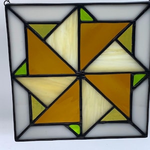 May include: A square stained glass panel featuring a geometric pinwheel pattern. The design consists of amber, cream, and bright lime green glass segments held together by dark metal solder lines, creating a symmetrical, multi-colored mosaic.