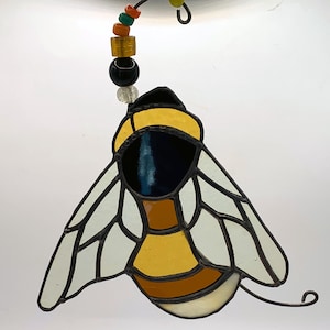 May include: Stained glass bee suncatcher with yellow, black, and white details. The bee has a yellow head and body with brown stripes, white wings, and a black outline. A beaded wire with colorful beads is attached to the top.