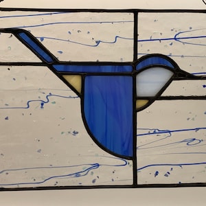 May include: Stained glass panel featuring a stylized blue bird in flight. The bird has a blue body and wings, with white and yellow accents. The background is white with blue swirls and speckles, framed by a black border.