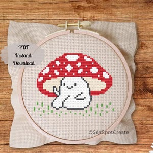 May include: A cross-stitch embroidery in a wooden hoop features a red and white mushroom with a smiling white character underneath. The text "PDF Instant Download" is visible. The design is on a beige fabric.