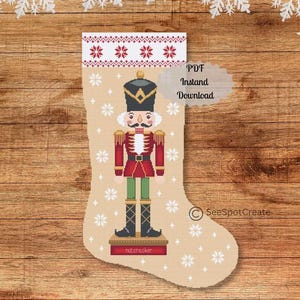 Puede incluir: Un calcetín navideño beige con un diseño de punto de cruz de un soldado cascanueces. El calcetín presenta un puño estampado en rojo y blanco y detalles de copos de nieve. El texto "PDF Instant Download" es visible.