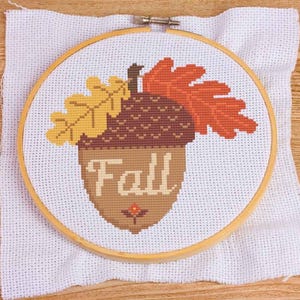 May include: A cross-stitch embroidery in a wooden hoop features a brown acorn with the word "FALL" and autumn leaves in yellow and orange. The embroidery is on white fabric, and the hoop is set against a wooden surface.