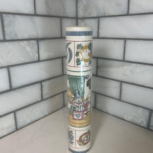 May include: A roll of decorative wallpaper featuring a repeating pattern of whimsical illustrations. The design includes a clown, floral elements, and other decorative motifs in a soft color palette of blue, green, and yellow. The wallpaper is rolled up and ready for use.