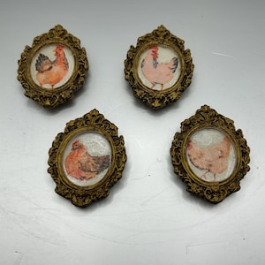 May include: Four small, ornate gold frames with a white background and a picture of a chicken in each frame. The chickens are all different colors and have different poses.