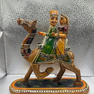 May include: A hand-painted wooden camel figurine with two figures riding on its back. The camel is brown with decorative painted details. The riders are dressed in colorful traditional clothing. The base is black with a decorative border.