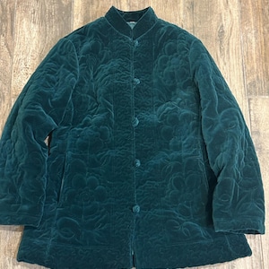 Puede incluir: Una chaqueta acolchada de color verde esmeralda oscuro con cuello mao y cuatro botones de palanca cubiertos de tela en el centro. La chaqueta está confeccionada en un material de terciopelo suave con un patrón floral en relieve por toda la tela.