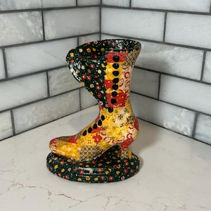 Könnte beinhalten: Eine dekorative Vase in Form eines Stiefels aus Keramik. Der Stiefel hat ein Patchwork-Design mit roten, gelben, grünen und schwarzen Blumenmustern. Er hat einen kleinen Absatz und eine Reihe schwarzer Knöpfe. Der Sockel ist ein rundes, grünes Blumenmuster.