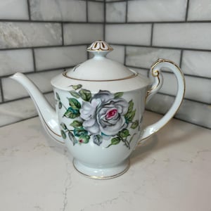 Könnte beinhalten: Weiße Porzellan-Teekanne mit Deckel, mit einem floralen Rosendesign in Grau-, Grün- und Rottönen. Die Teekanne hat eine goldene Verzierung und einen Griff sowie einen Ausguss zum Ausgießen. Ein dekoratives Stück zum Servieren von Tee.