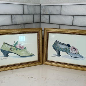 May include: Two framed watercolor paintings of vintage shoes. One shoe is light green with a dark blue heel and a pink flower. The other is blue-grey with a black heel and a pink flower. Both are in gold frames.