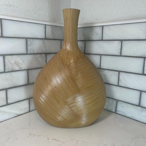 Puede incluir: Un jarrón decorativo alto de color beige claro con un patrón texturizado similar al tejido. El jarrón tiene una base redondeada y un cuello largo y delgado. El diseño presenta líneas diagonales que crean un efecto geométrico. El jarrón se exhibe sobre una encimera blanca.