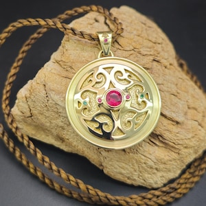 May include: A gold-colored pendant with a circular design, featuring a central red gemstone and smaller green and red accents. The pendant hangs from a brown braided cord. The design has an intricate, openwork pattern.