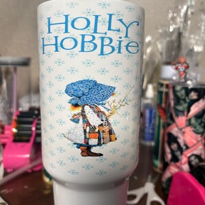 May include: White tumbler featuring the text "HOLLY HOBBIE" in blue, with a floral pattern and a graphic of a girl in a patchwork dress and bonnet. The tumbler has a white base and a cylindrical body.