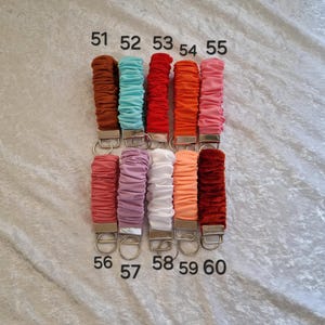 May include: Ten fabric-covered keychains with metal hardware, arranged in two rows of five. Each keychain features a gathered, scrunchie-style textile in colors including brown, teal, red, orange, pink, mauve, white, peach, and dark red, labeled 51-60.