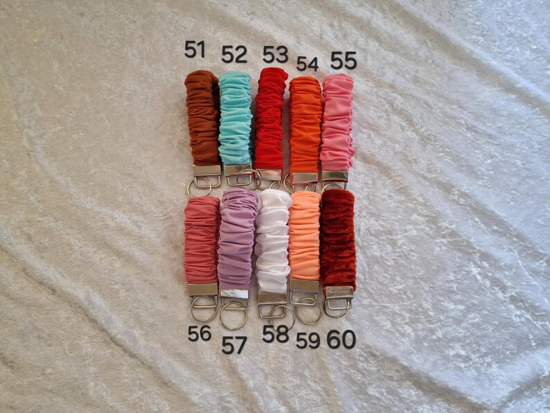 May include: Ten fabric-covered keychains with metal hardware, arranged in two rows of five. Each keychain features a gathered, scrunchie-style textile in colors including brown, teal, red, orange, pink, mauve, white, peach, and dark red, labeled 51-60.