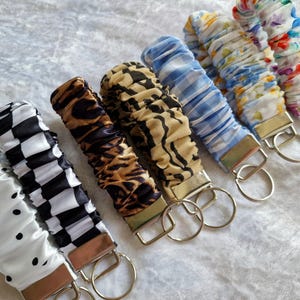 May include: A collection of seven fabric wristlet keychains featuring various patterns including white with black dots, black and white checkerboard, brown leopard print, tan and black zebra print, blue and white gingham, floral, and colorful abstract prints.