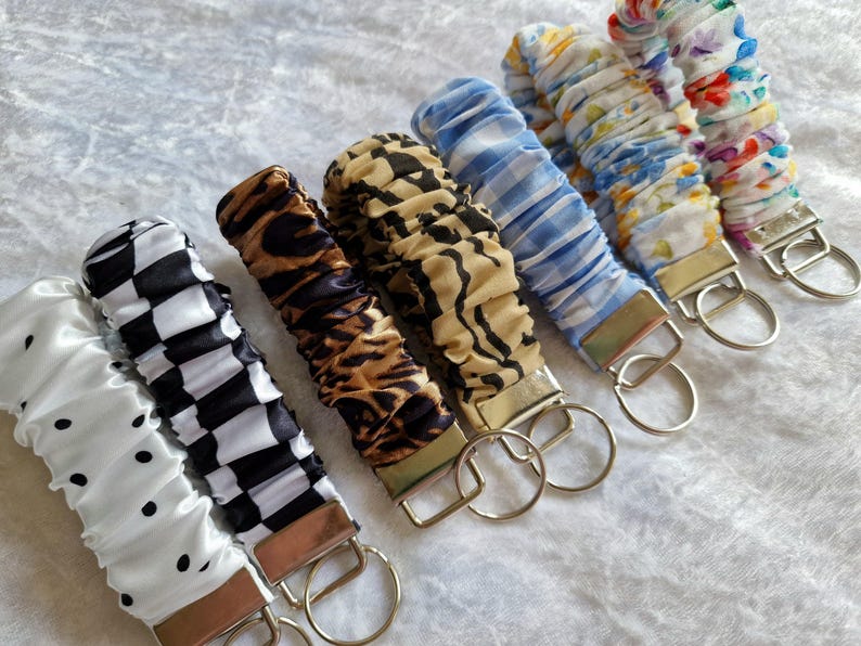 May include: A collection of seven fabric wristlet keychains featuring various patterns including white with black dots, black and white checkerboard, brown leopard print, tan and black zebra print, blue and white gingham, floral, and colorful abstract prints.